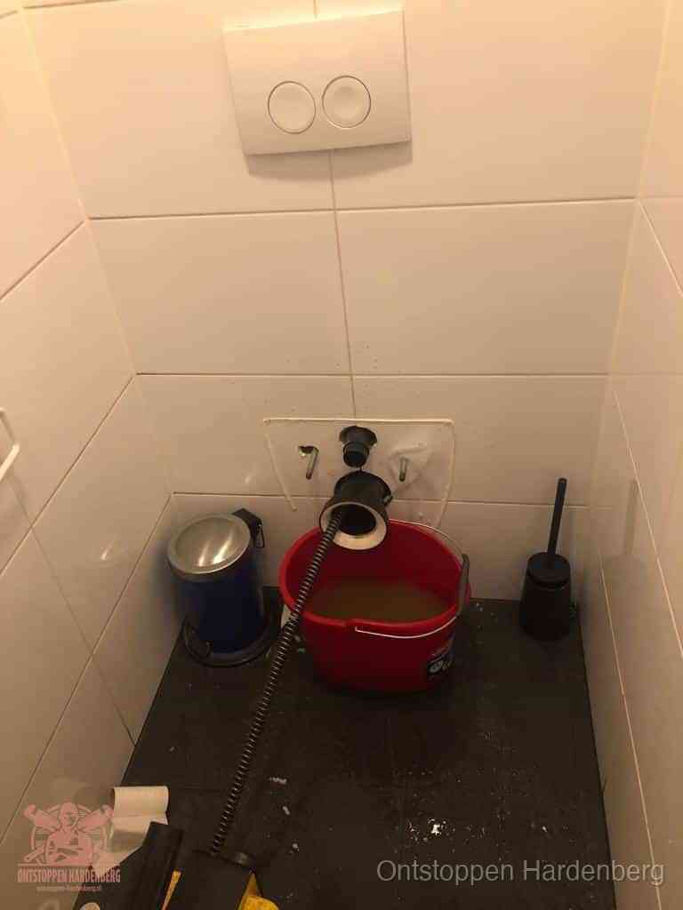 Een ontstopte wand-wc met verwijderde toiletpot waar een ontstoppingsveer in de wanduitgang verdwijnt, het achterhalen waar de verstopping zit in Hardenberg