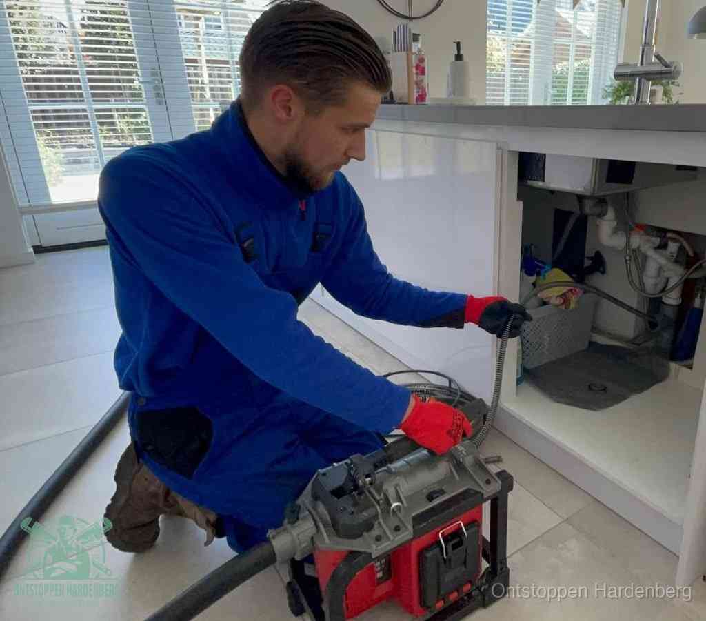 Een monteur met rode handschoenen knielt in een keuken en voert een elektrische ontstoppingsveer in tijdens het professioneel afvoer ontstoppen in Hardenberg